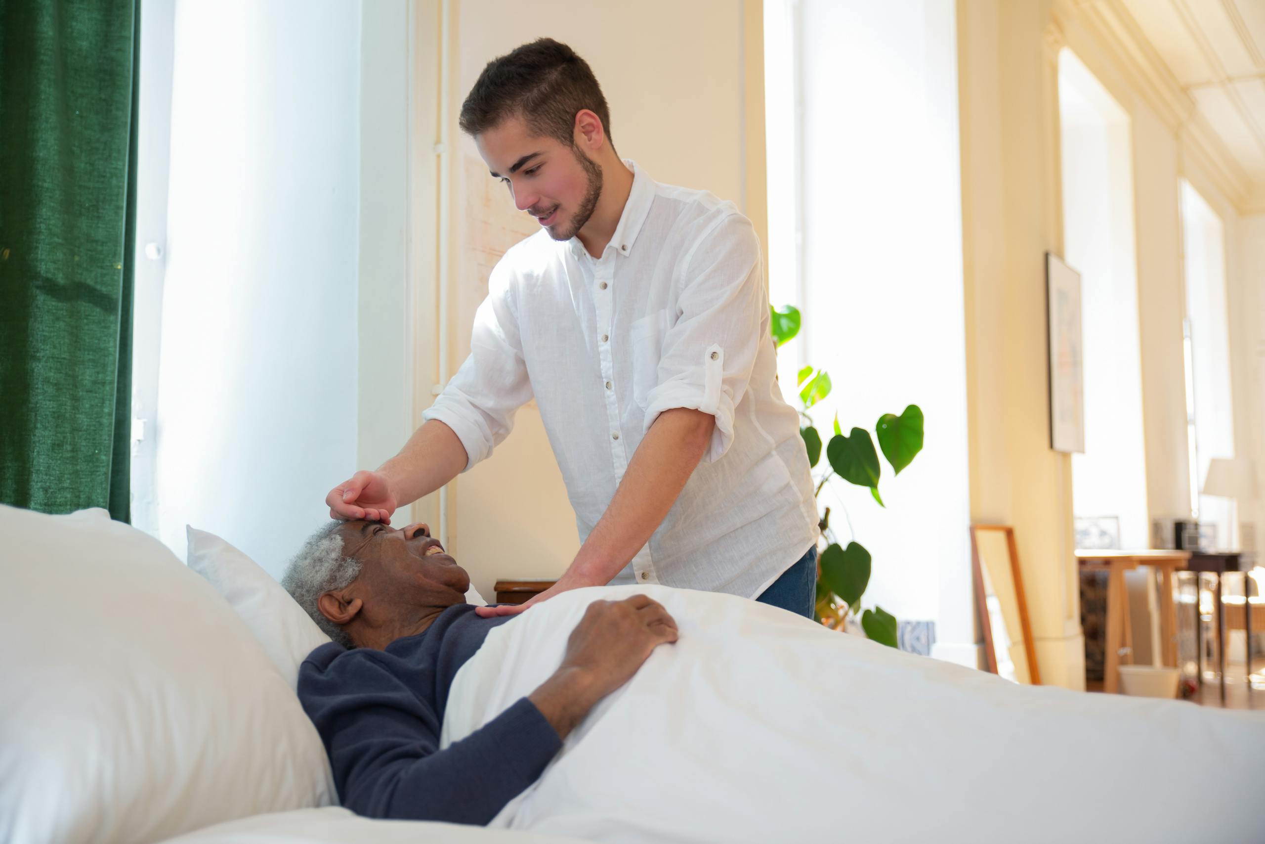 A Man in White Shirt Standing Beside an Elderly Lying on the Bed